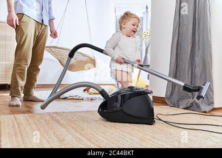 Niedlicher Baby Junge hält Staubsauger, während er zu Hause auf dem Teppich steht Stockfoto