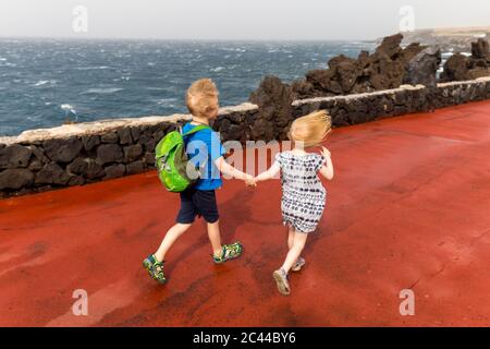 Hochwinkelansicht von Geschwistern, die sich beim Laufen auf dem Pier auf dem Meer an der Costa Adeje, Kanarische Inseln, Spanien, die Hände halten Stockfoto