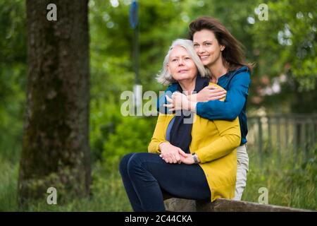 Porträt einer glücklichen Frau, die ihre Mutter umarmt, die auf der Rückenlehne einer Parkbank sitzt Stockfoto