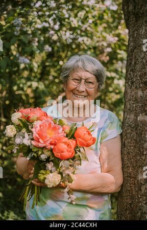 Lächelnde ältere Frau mit frischem Pfingstrosen-Bouquet am Baumstamm im Garten Stockfoto