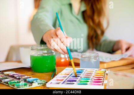 Junge Frau taucht Pinsel in Aquarell auf Tisch zu Hause Stockfoto