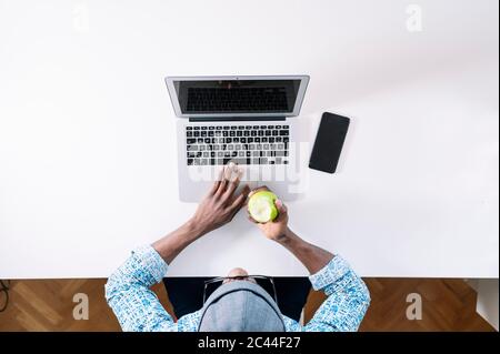 Junger Mann, der grünen Apfel isst, während er am Schreibtisch zu Hause arbeitet Stockfoto