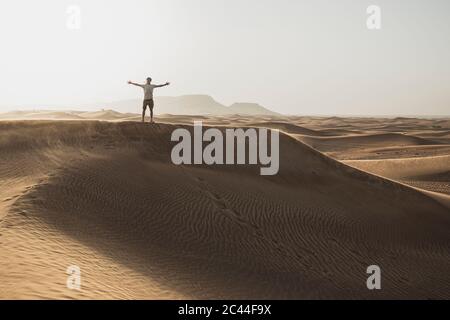 Mittlere Entfernung von männlichen Touristen mit Armen ausgestreckt auf Sanddünen in der Wüste in Dubai, Vereinigte Arabische Emirate Stockfoto