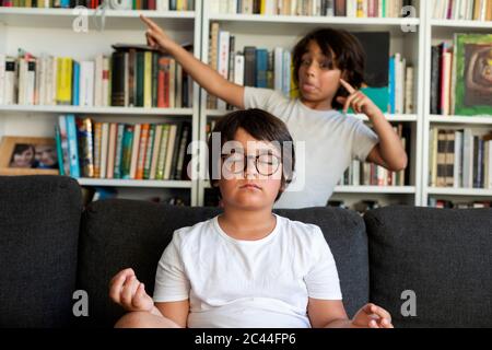 Junge auf der Couch sitzend meditierend, sein Bruder neckend ihn Stockfoto