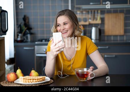 Glückliche Frau mit Smartphone und Ohrstöpseln sitzen am Küchentisch zu Hause Stockfoto