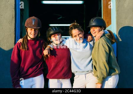 Porträt von fröhlichen weiblichen Jockeys stehen zusammen gegen Stall an sonnigen Tag Stockfoto