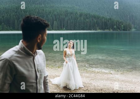 Schöne junge kasachische Braut in einem weißen Kleid ist auf dem Hintergrund des Sees zu Fuß. Der Bräutigam schaut sie an. Stockfoto