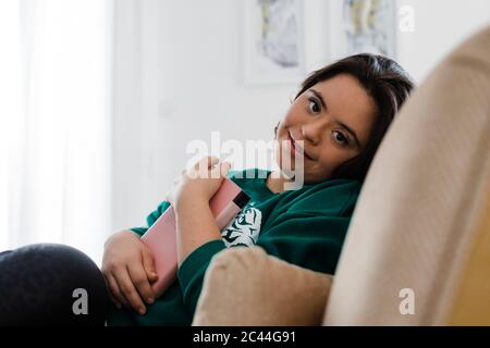 Lächelnde junge Frau mit Down-Syndrom hält Buch, während sie sich auf dem Sessel zu Hause Stockfoto