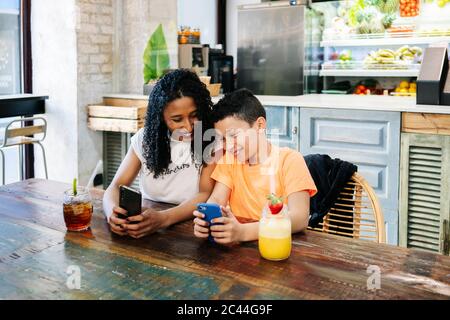 Fröhliche Mutter und Sohn mit Handys am Tisch im Restaurant Stockfoto