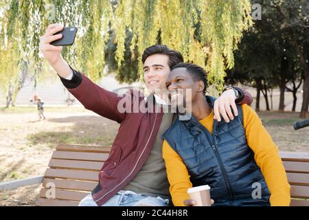 Zwei glückliche junge Männer sitzen auf Parkbank und machen ein Selfie Stockfoto