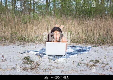 Junge Frau, die mit einem Laptop auf der Decke am Strand liegt Stockfoto