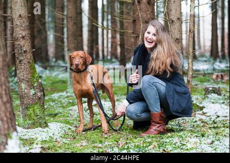 Lächelnde junge schöne Frau mit langen braunen Haaren, die vom Hund gegen Bäume im Wald hocken Stockfoto