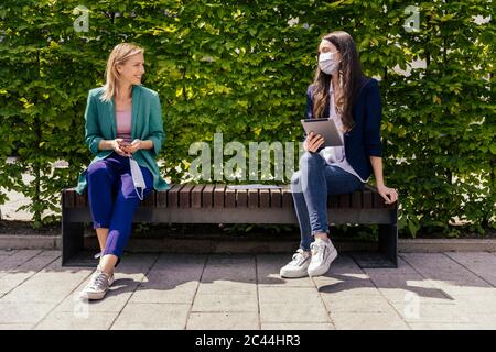 Zwei Geschäftsfrauen sitzen auf der Bank draußen und halten ihre Distanz, während sie Gesichtsmaske tragen Stockfoto