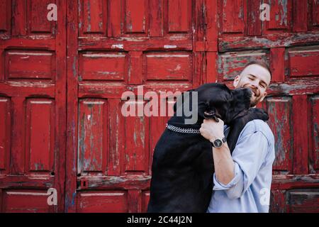 Porträt eines lachenden jungen Mannes und seines Hundes vor einer alten roten Holztür Stockfoto