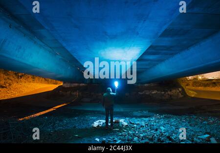 Spanien, Galicien, Rückansicht eines Mannes mit Kapuze, der unter einer Betonbrücke mit hellblauem Licht in der Hand steht Stockfoto