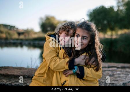 Junge umarmende lächelnde Schwester in gelben Regenmänteln am Flussufer Stockfoto