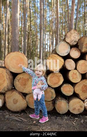 Portrait von glücklichen kleinen Mädchen mit Puppe vor Stapel Holz im Wald stehen Stockfoto