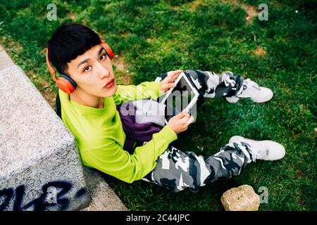 Junger Mann mit Kopfhörern mit digitalem Tablet, während er auf Gras sitzt Stockfoto