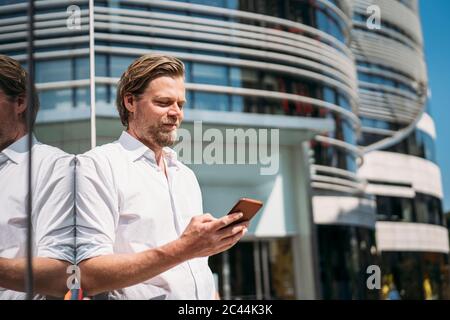 Geschäftsmann in der Stadt lehnt sich an Glasfront Überprüfung Smartphone Stockfoto
