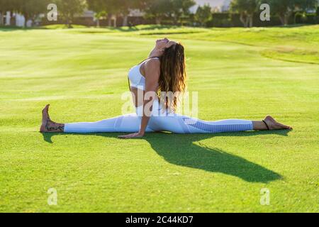 Frau, die Yoga auf Rasen bei Sonnenschein praktiziert, dabei die Splits macht Stockfoto