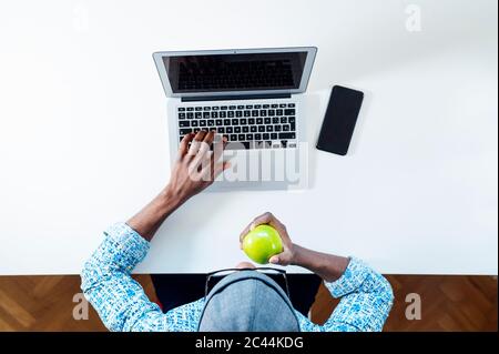 Junger Mann, der grünen Apfel isst, während er zu Hause arbeitet Stockfoto