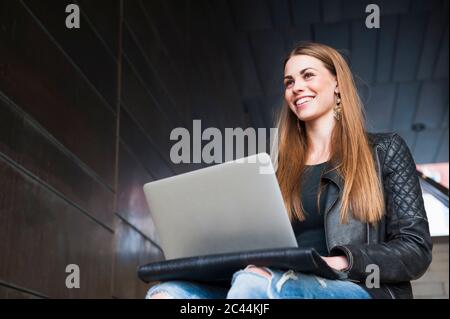 Lächelnde junge Frau mit Laptop, während sie in einem unterirdischen Gang sitzt Stockfoto