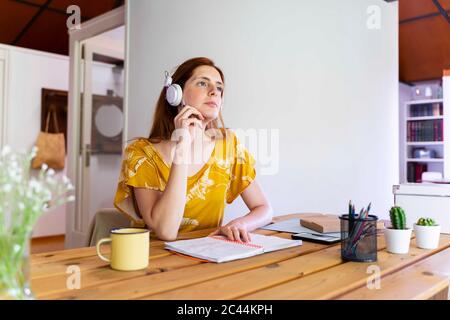 Eine durchdachte Frau mit Kopfhörern sitzt am Schreibtisch im Haus Stockfoto