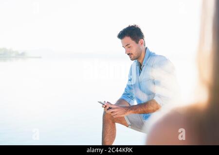 Porträt eines jungen Mannes, der vor einem See steht und auf das Handy schaut Stockfoto