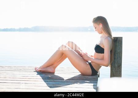 Junge Frau sitzt auf dem Steg vor einem See und hört Musik mit Smartphone und Kopfhörer Stockfoto