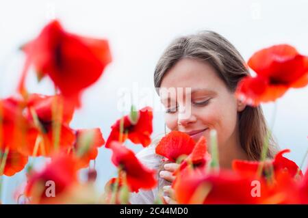 Lächelnde Frau riecht frische rote Mohnblume gegen den Himmel Stockfoto