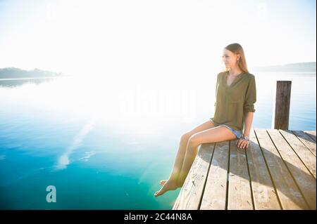 Junge Frau, die bei Sonnenaufgang am Anlegesteg am See sitzt Stockfoto