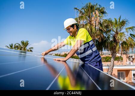 Lächelnder reifer Techniker, der Solarpanel auf dem Hausdach gegen den blauen Himmel installiert Stockfoto