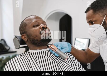 Friseur trägt chirurgische Maske und Handschuhe schneiden Bart des Kunden Stockfoto