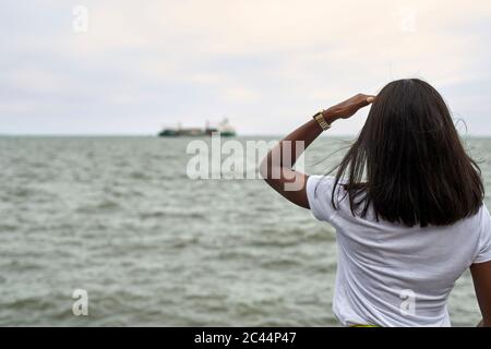 Rückansicht einer jungen Frau vor dem Meer, die am Horizont auf das Schiff schaut Stockfoto