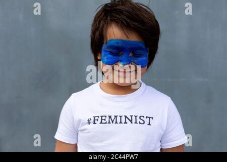 Porträt eines lächelnden Jungen mit bemalter blauer Maske auf seinem Gesicht in T-Shirt mit Aufdruck 'Feminist' Stockfoto