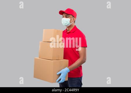 Lieferung Mann Holding Karton Boxen Tragen medizinische Maske und Handschuhe Beobachtungsseite isoliert. Indian Delivery Boy lächelnd mit Boxen in den Händen Stockfoto