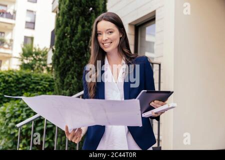 Portrait einer lächelnden Frau mit digitalem Tablet und Gesichtsmaske in der Hand vor einem Haus Stockfoto