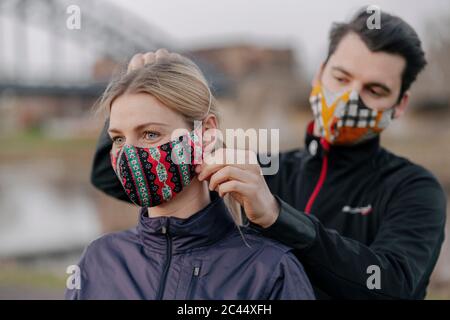 Junger Mann, der während der COVID-19 im Park steht, die Gesichtsmaske für die Frau anpasst Stockfoto