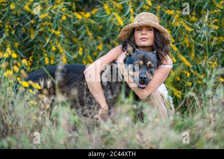 Porträt einer schönen jungen Frau, die Hund umarmt, während sie zwischen Pflanzen hockend, Alicante, Provinz Alicante, Spanien Stockfoto