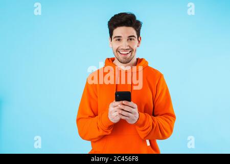 Porträt eines fröhlichen jungen Mannes mit Hoodie, isoliert auf blauem Hintergrund stehend, Mobiltelefon haltend Stockfoto