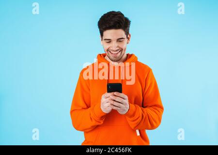 Porträt eines fröhlichen jungen Mannes mit Hoodie, isoliert auf blauem Hintergrund stehend, Mobiltelefon haltend Stockfoto