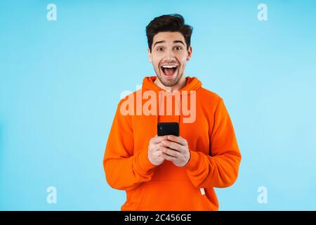 Porträt eines fröhlichen jungen Mannes mit Hoodie, isoliert auf blauem Hintergrund stehend, Mobiltelefon haltend Stockfoto