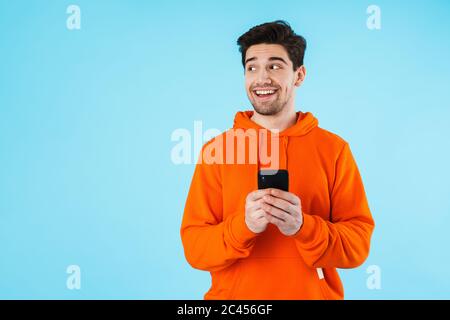 Porträt eines fröhlichen jungen Mannes mit Hoodie, der isoliert auf blauem Hintergrund steht, das Mobiltelefon hält und wegschaut Stockfoto