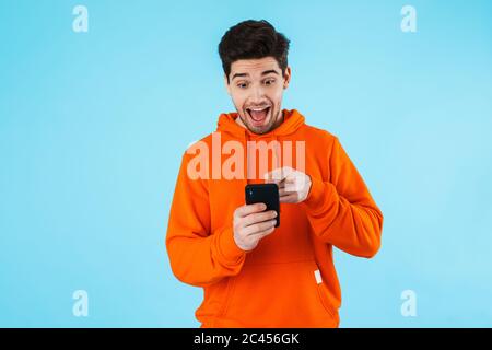 Porträt eines fröhlichen jungen Mannes mit Hoodie, isoliert auf blauem Hintergrund stehend, Mobiltelefon haltend Stockfoto