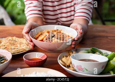 Frau Hände hält eine Schüssel mit thailändischen lokalen hausgemachten Speisen roten Curry mit Huhn in Keramikschale am Esstisch Stockfoto