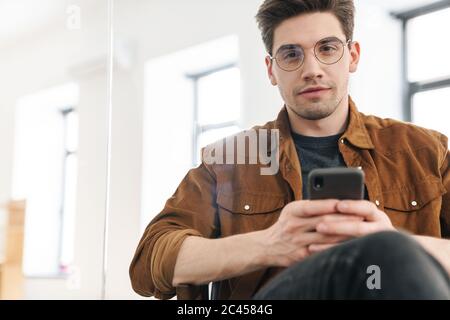 Bild von gutaussehenden selbstbewussten jungen Mann in Brillen mit Handy während der Arbeit im Büro Stockfoto