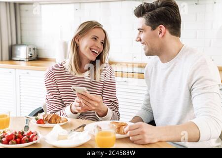 Portrait des jungen glücklichen Paares mit Smartphone beim Frühstück in der gemütlichen Küche zu Hause Stockfoto