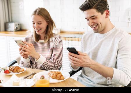 Porträt eines jungen lächelnden Paares mit Smartphones beim Frühstück in der gemütlichen Küche zu Hause Stockfoto