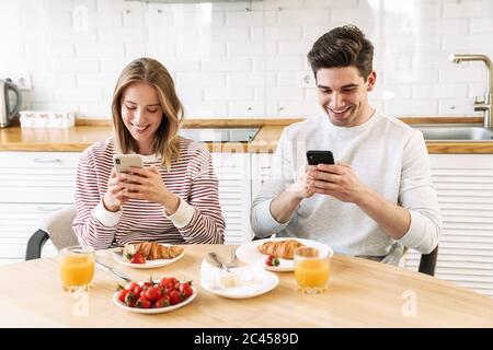 Porträt eines jungen lächelnden Paares mit Smartphones beim Frühstück in der gemütlichen Küche zu Hause Stockfoto