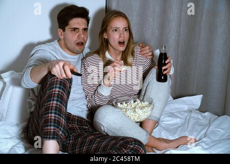 Porträt eines schockierten kaukasischen Paares Fernsehen und Popcorn essen, während sie zu Hause im Bett sitzen Stockfoto